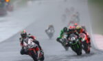 Motorcycle racer leading a wet track race, leaning into a turn with rain-slick pavement and a pack of riders behind him on colorful bikes.