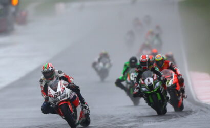 Motorcycle racer leading a wet track race, leaning into a turn with rain-slick pavement and a pack of riders behind him on colorful bikes.