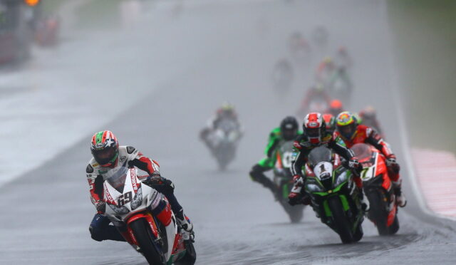 Motorcycle racer leading a wet track race, leaning into a turn with rain-slick pavement and a pack of riders behind him on colorful bikes.