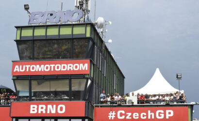 Control tower at Brno Automodrom with bold red signs 'AUTOMODROM' and 'BRNO', spectators on the balcony, and a white event tent nearby.