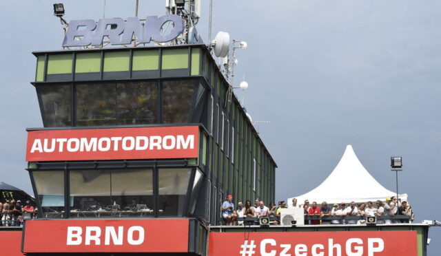 Control tower at Brno Automodrom with bold red signs 'AUTOMODROM' and 'BRNO', spectators on the balcony, and a white event tent nearby.