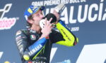 MotoGP rider in a Yamaha suit kisses a trophy on a podium, with a blue backdrop and sponsor banners behind him.