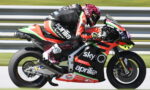 MotoGP rider on a red-and-black Aprilia racing bike with Sky Q branding speeding along a track, leaning into a turn. Non-visual alternative: racing scene with sponsor logos on bike and suit, motorcycle in motion on race track.