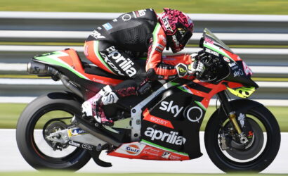 MotoGP rider on a red-and-black Aprilia racing bike with Sky Q branding speeding along a track, leaning into a turn. Non-visual alternative: racing scene with sponsor logos on bike and suit, motorcycle in motion on race track.