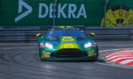 Green race car with bright yellow accents and sponsor logos speeding through a curving race track, barriers and a large Debka banner in the background