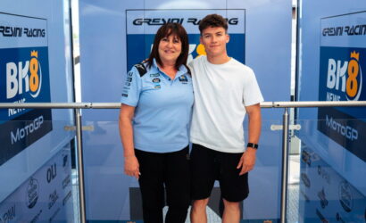 Woman in a light blue polo with sponsor logos stands beside a young man in a white t-shirt; both pose in front of Greyni Racing banners at a event backdrop.