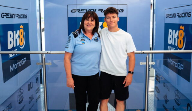 Woman in a light blue polo with sponsor logos stands beside a young man in a white t-shirt; both pose in front of Greyni Racing banners at a event backdrop.