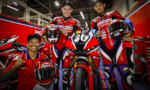 Four riders in red Honda racing suits posing with a red race bike numbered 30 in a garage/pit area, smiling and giving a thumbs-up.