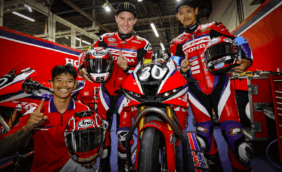 Four riders in red Honda racing suits posing with a red race bike numbered 30 in a garage/pit area, smiling and giving a thumbs-up.
