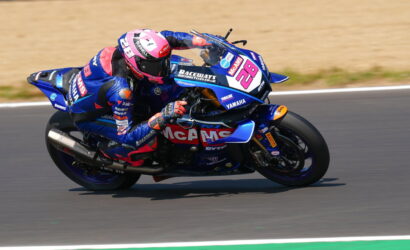 Moto racer in blue-pink livery leaning through a sharp turn on a numbered Yamaha race bike (No. 28) with McAMS branding, on a track surface.