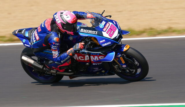 Moto racer in blue-pink livery leaning through a sharp turn on a numbered Yamaha race bike (No. 28) with McAMS branding, on a track surface.