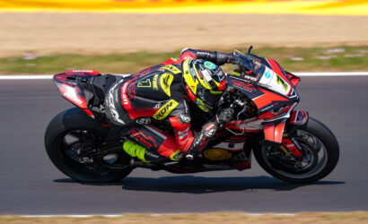 Motorcycle racer in red and black suit leaning into a high-speed bend on a racing track with sponsor graphics on the bike's fairings
