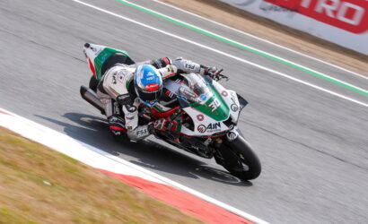 Motorcycle racer in white, green, and red suit leans into a sharp turn on a racetrack, bike number 30 close to the ground.