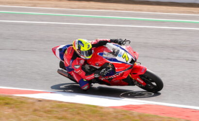 Motorcycle racer in red gear leaning into a turn on a race track, riding a red Honda bike with blue and white accents.