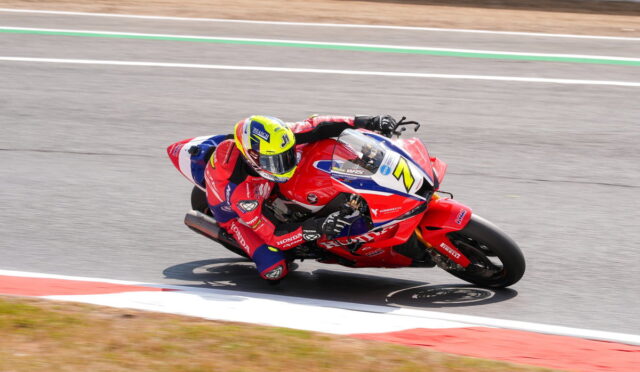 Motorcycle racer in red gear leaning into a turn on a race track, riding a red Honda bike with blue and white accents.