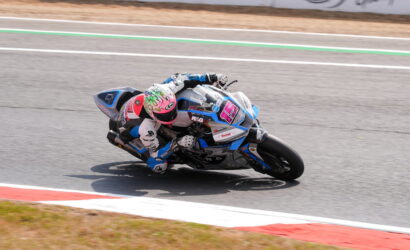 Motorcycle racer leaning into a sharp turn on a race track, wearing white and blue gear with a pink helmet.