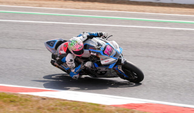 Motorcycle racer leaning into a sharp turn on a race track, wearing white and blue gear with a pink helmet.