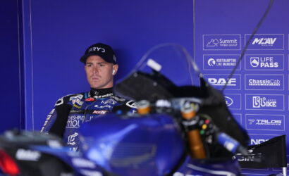 Moto racer in racing suit sits in a blue, sponsor‑stamped backdrop, wearing AirPods and a cap, with a blue motorcycle blurred in foreground.