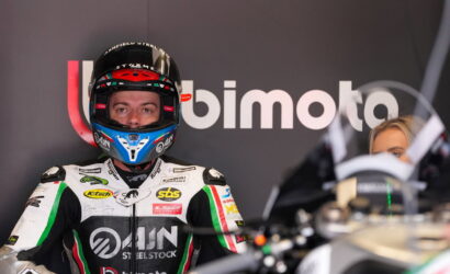 Motorcycle racer in full white-green-black suit and blue helmet stands in front of a BIMOTA backdrop.