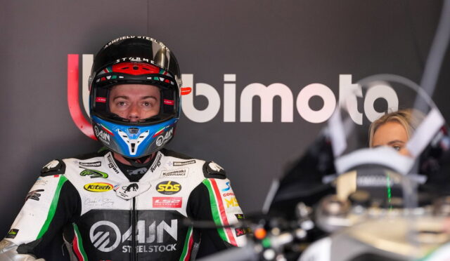Motorcycle racer in full white-green-black suit and blue helmet stands in front of a BIMOTA backdrop.