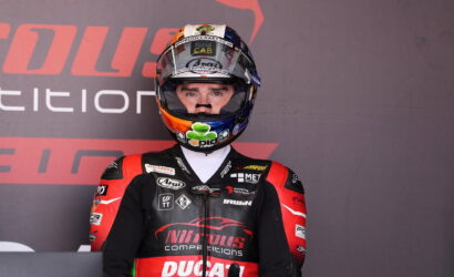 Motorcycle racer in red-and-black Ducati leathers wearing a full-face helmet in a pit area backdrop with sponsor logos.