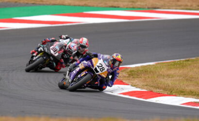Motorcycle racers lean into a sharp corner on a track with red-and-white curbs; rider #28 leads in purple gear.