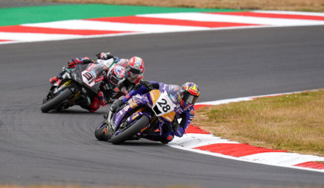 Motorcycle racers lean into a sharp corner on a track with red-and-white curbs; rider #28 leads in purple gear.