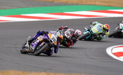Moto racers lean into a curve on a race track; purple bike #28 leads the pack, closely followed by others in colorful leathers and helmets on the tarmac.