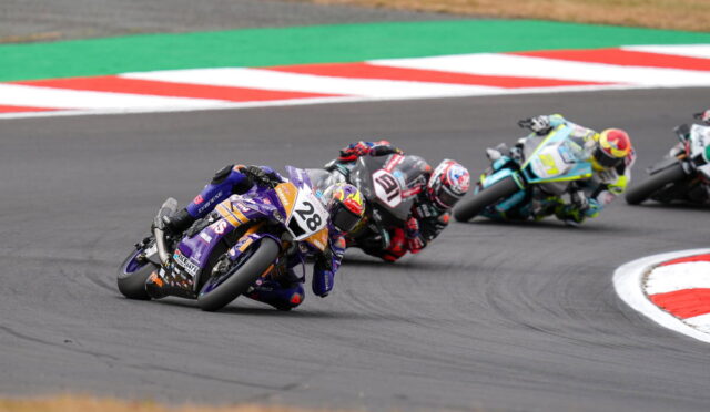 Moto racers lean into a curve on a race track; purple bike #28 leads the pack, closely followed by others in colorful leathers and helmets on the tarmac.