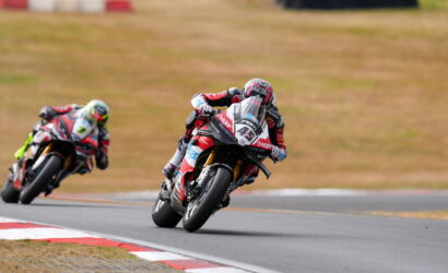 A motorcycle racer in red and white gear leans into a turn on a race track, with a second rider close behind on the left.
