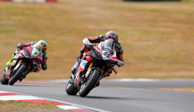 A motorcycle racer in red and white gear leans into a turn on a race track, with a second rider close behind on the left.