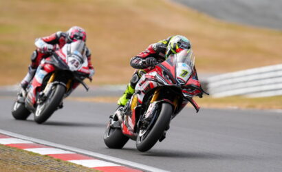 Two motorcycle racers lean into a sharp turn on a race track, front rider in red/white gear and neon yellow accents.