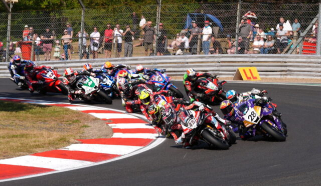 Group of motorcycle racers lean into a tight turn on a race track, closely following each other as spectators watch from behind a fence.
