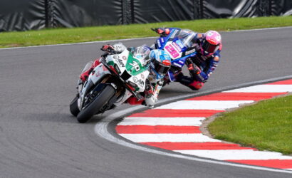 Two motorcycle racers lean into a sharp corner on a racetrack, red-and-white curbs visible and grass beyond.