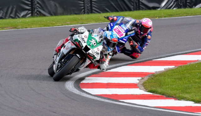 Two motorcycle racers lean into a sharp corner on a racetrack, red-and-white curbs visible and grass beyond.