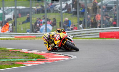 Rider in yellow/orange gear and a helmet leans into a sharp left turn on a race track, number 89 on the bike.