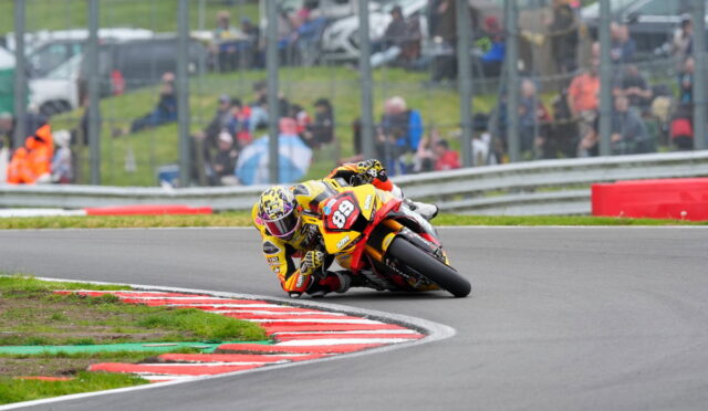 Rider in yellow/orange gear and a helmet leans into a sharp left turn on a race track, number 89 on the bike.
