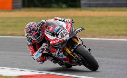 Professional motorcycle racer in red-and-white livery leans deeply into a sharp turn on a race track, helmeted and gripping the bike.