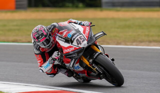 Motorcycle racer in red-and-white racing suit leans into a sharp turn on a track, sponsor decals visible on bike and gear