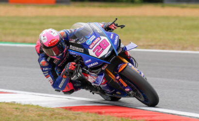 Motorcycle racer in blue and pink gear leaning into a turn on a blue Yamaha race bike with number 28 on the fairing.