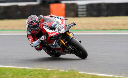 Racing motorcycle rider in red and white gear leans hard into a left-hand turn on a race track, helmet visors and sponsor logos visible