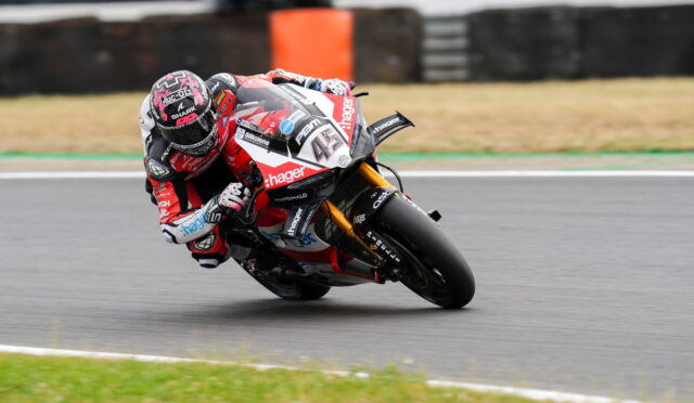 Racing motorcycle rider in red and white gear leans hard into a left-hand turn on a race track, helmet visors and sponsor logos visible