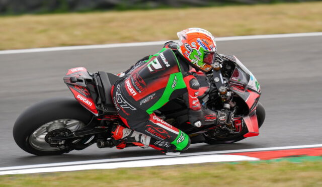 Motorcycle racer in red-green suit leaning into a curve on a race track, Ducati branding visible on bike and suit