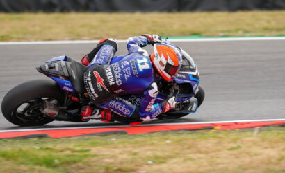 MotoGP rider in a blue suit and orange helmet leans into a curve on a track, blue Yamaha bike with sponsor decals visible.