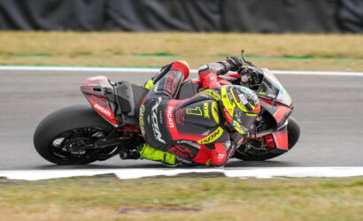 MotoGP rider in red/black leathers leaning into a wide corner on a red Ducati bike with bright yellow helmet.