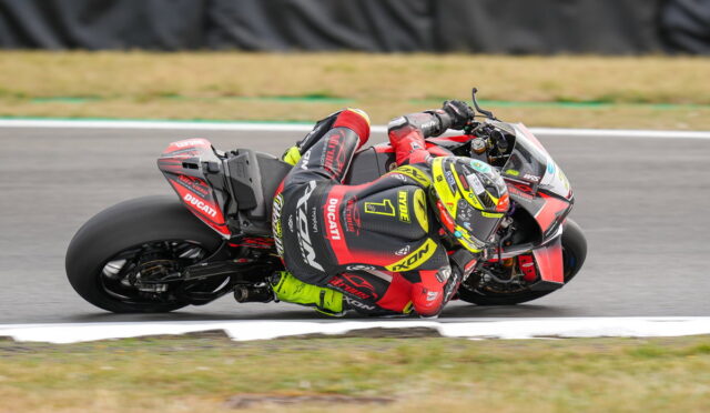 MotoGP rider in red/black leathers leaning into a wide corner on a red Ducati bike with bright yellow helmet.