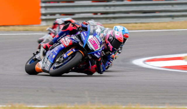 Rider in pink helmet on blue Yamaha bike leans into a sharp turn, racing closely with another rider behind them on a track.