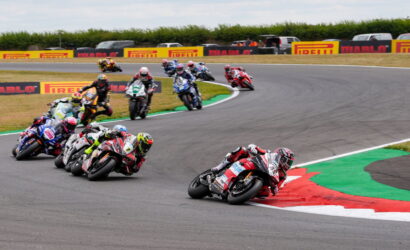 Group of motorcycle racers leaning into a sharp track bend, close together on a racing circuit with Pirelli banners in the background.