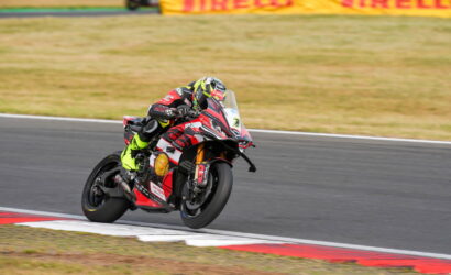 Motorcycle racer in neon-yellow suit leaning into a corner on a race track with a red-and-white bike in the foreground.