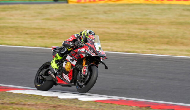Motorcycle racer in neon-yellow suit leaning into a corner on a race track with a red-and-white bike in the foreground.